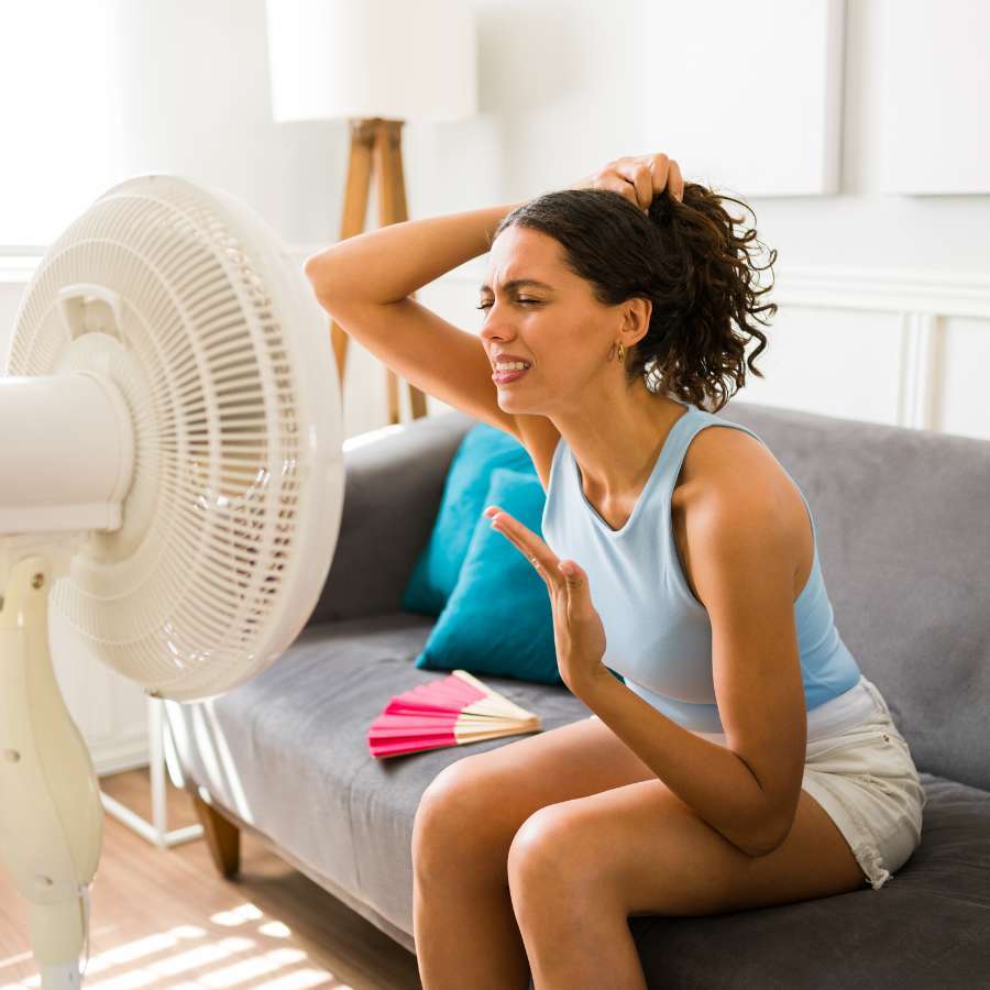 Woman sitting in front of a fan holding her hair back and wincing at the heat.
